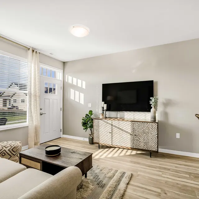 A modern living room featuring a beige sofa, a wooden coffee table, and a patterned sideboard. Natural light enters through large windows with white blinds. A television mounted on the wall and a door leading outside adds to the decor.