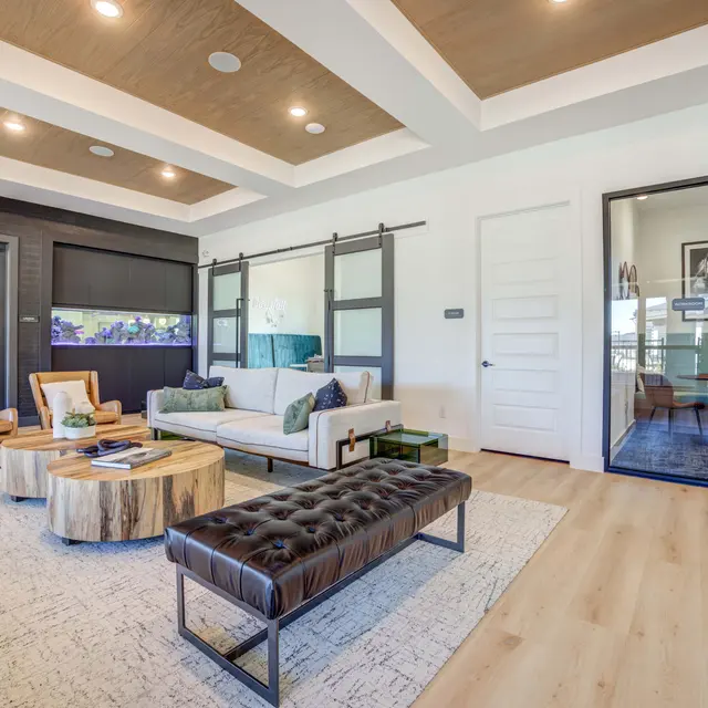 A modern living room featuring a white sofa with blue and beige cushions, a large round wooden coffee table, a tufted leather ottoman, and two wooden armchairs. The ceiling has a coffered design, and glass sliding doors lead to a separate room.