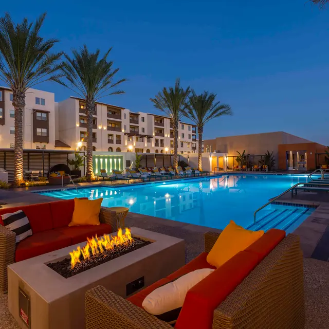 Evening Luxury Pool Area An outdoor pool area with seating around a fire pit, palm trees, and modern buildings in the background at dusk.