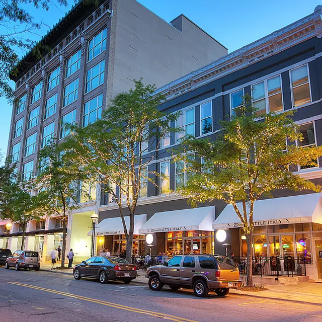 A city street at dusk with trees and buildings. One side features restaurant facades and outdoor seating. Cars are parked along the curb.