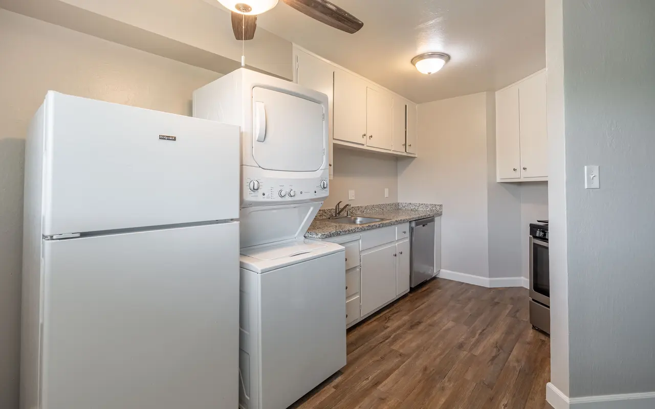 A modern kitchen featuring a white refrigerator, stacked washer and dryer, and a stove, with light wood flooring and gray walls.