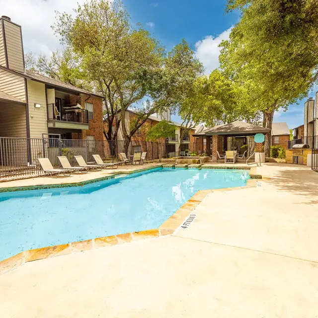 A clear swimming pool surrounded by lounge chairs and green trees, with apartment buildings in the background.