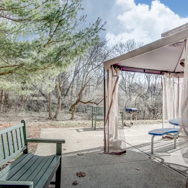 A backyard patio area featuring a green wooden bench and a gazebo with sheer curtains, surrounded by trees and a concrete floor.