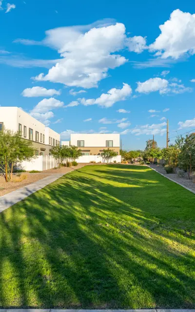 Urban Green Pathway A well-maintained green pathway surrounded by trees, leading to modern buildings under a blue sky with scattered clouds.