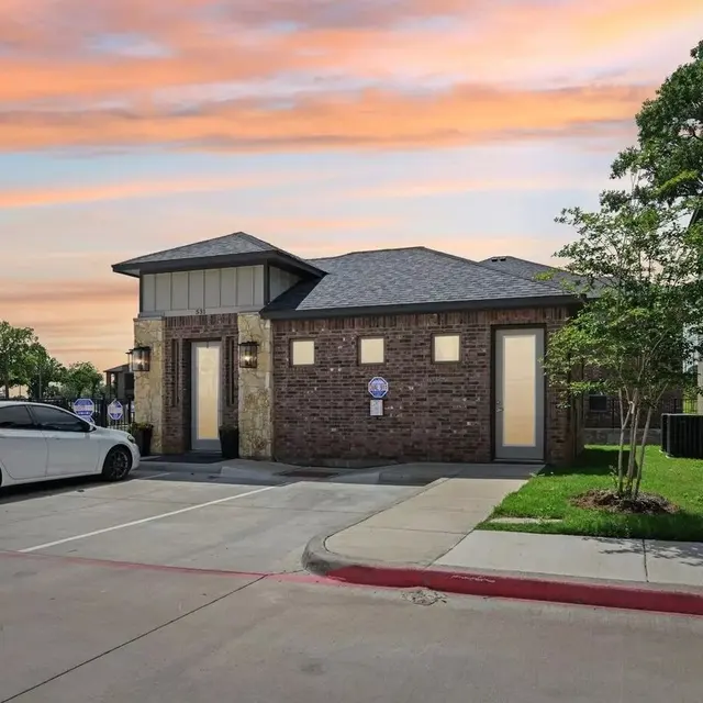 A sleek, modern building with a brick exterior and a slate roof, adjacent to a parking lot with a white car. The background features a vibrant sunset sky with shades of orange and pink, and green trees in the distance.