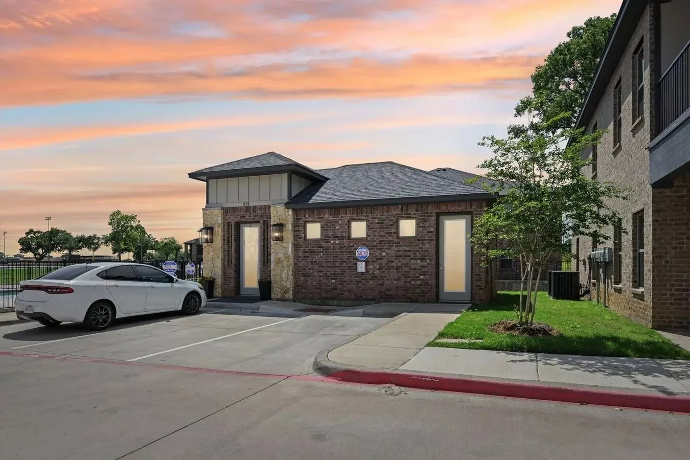 Modern Building Near Parking Lot at Sunset A sleek, modern building with a brick exterior and a slate roof, adjacent to a parking lot with a white car. The background features a vibrant sunset sky with shades of orange and pink, and green trees in the distance.