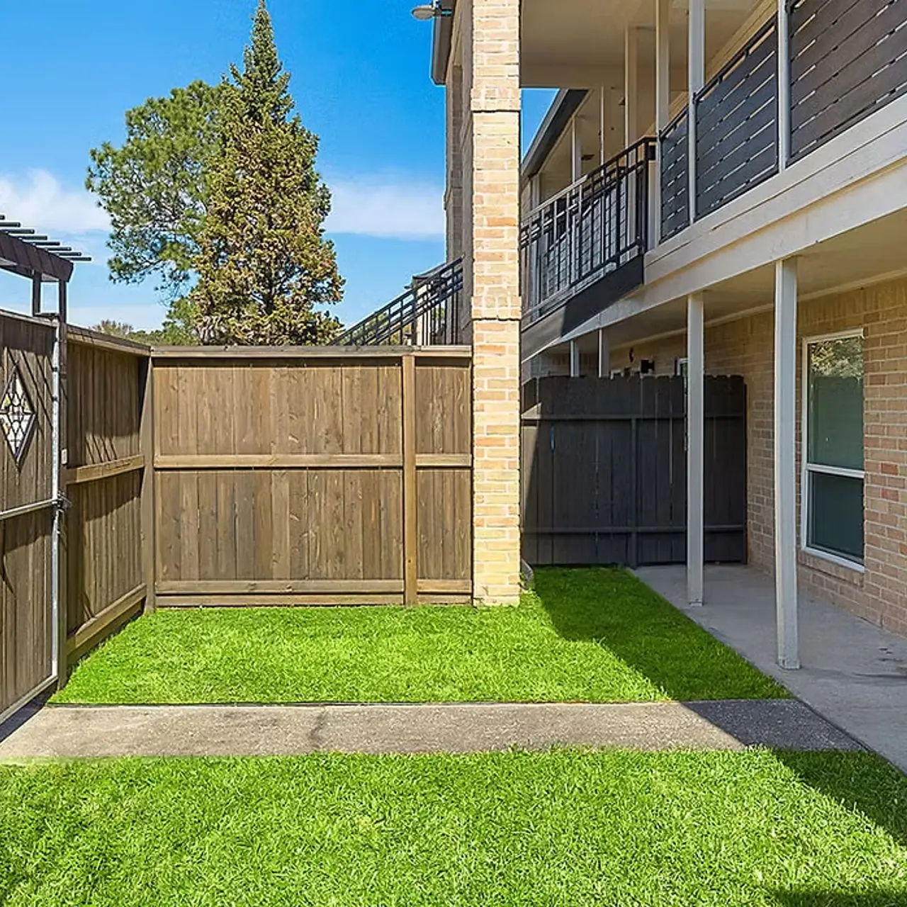 A view of a landscaped area between apartment buildings, featuring green grass and a wooden fence.