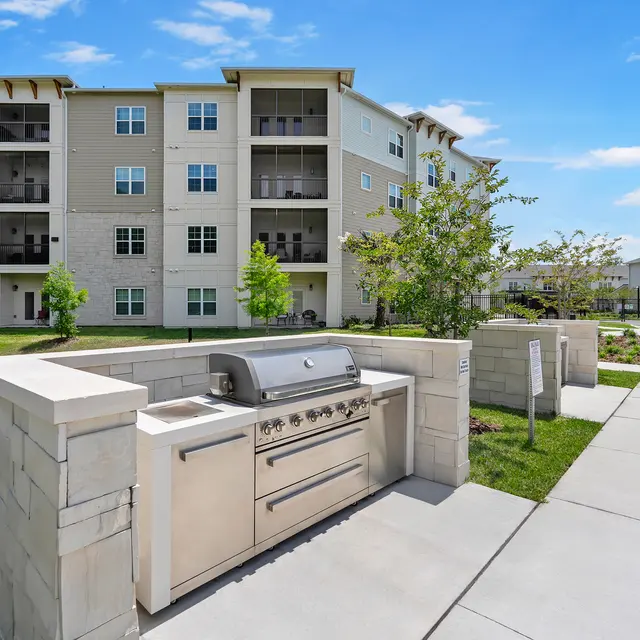 Outdoor Grill Area Near Apartment Complex A modern outdoor grill area in front of a multi-story apartment building with landscaped greenery.
