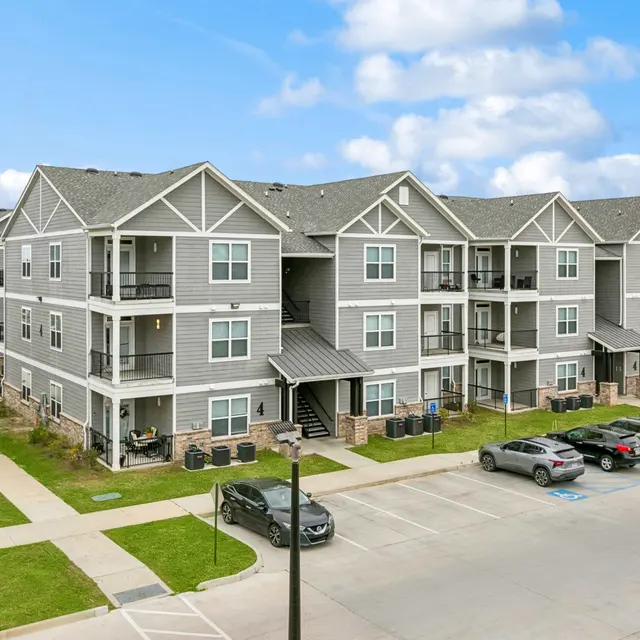 A modern multi-story apartment complex with gray siding and multiple balconies, surrounded by green grass and parking spaces.