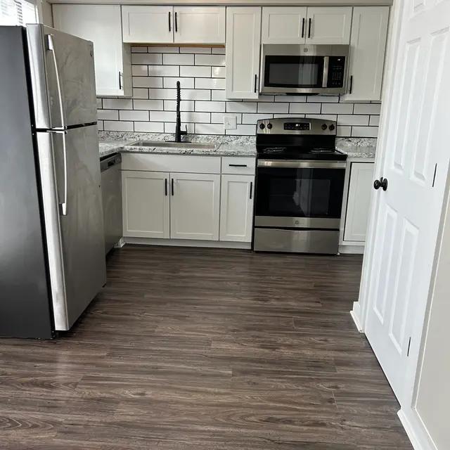 A modern kitchen featuring stainless steel appliances, including a refrigerator, microwave, and stove. The cabinets are white, and the backsplash is a stylish arrangement of white subway tiles. The flooring is a dark wood laminate, and there is significant natural light coming through the windows.