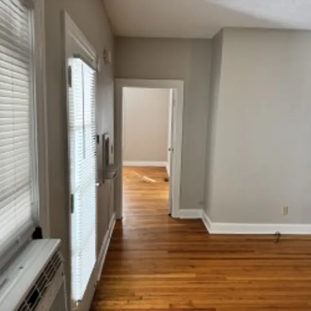 A view of an empty room with hardwood floors, featuring a door leading to another room and a window with blinds.
