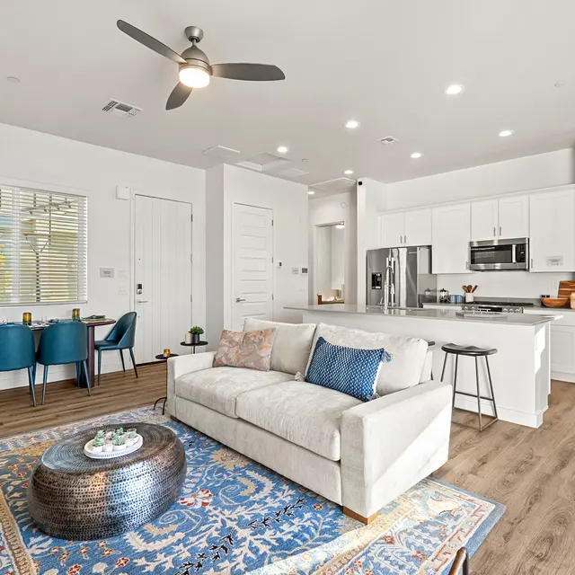 Modern Apartment Living Room A modern living space featuring a light-colored sofa, a round coffee table, and decorative elements. The room has a sleek kitchen visible in the background with stainless steel appliances. There are blue dining chairs set around a table, and the flooring is wood-like.