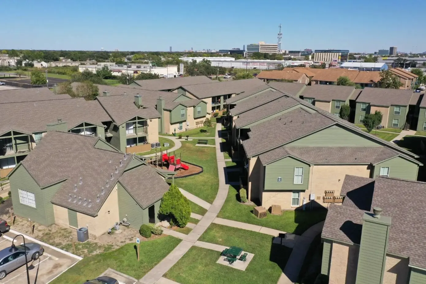 Aerial view of an apartment complex with green buildings and pathways. There is a playground area visible in the center.