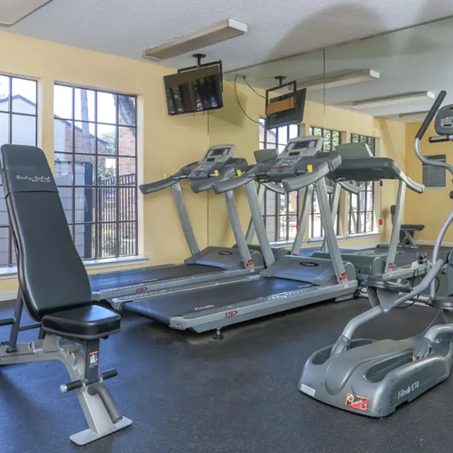 A fitness center with various exercise equipment including treadmills, an elliptical machine, and a weight bench under natural light from large windows.