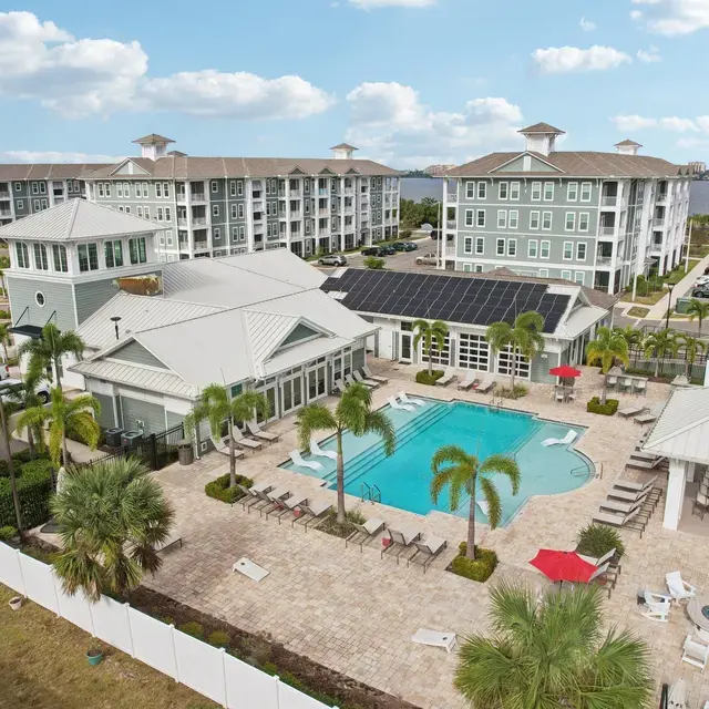 Aerial view of an apartment complex featuring a swimming pool surrounded by lounge chairs and palm trees, with additional buildings in the background and a lake visible.