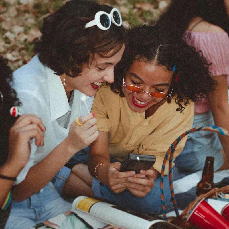 Friends Enjoying a Day Out Two young women laughing together while looking at a phone, surrounded by snacks and magazines in an outdoor setting.