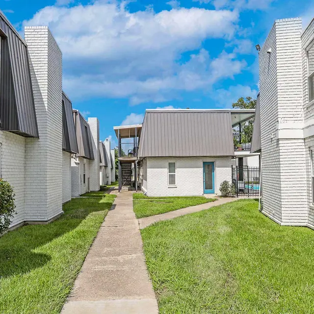 A well-maintained pathway between white brick apartment buildings with sloped roofs, featuring green grass and blue sky with clouds above. The pathway leads to a building with a blue door and a wooden deck in the background.