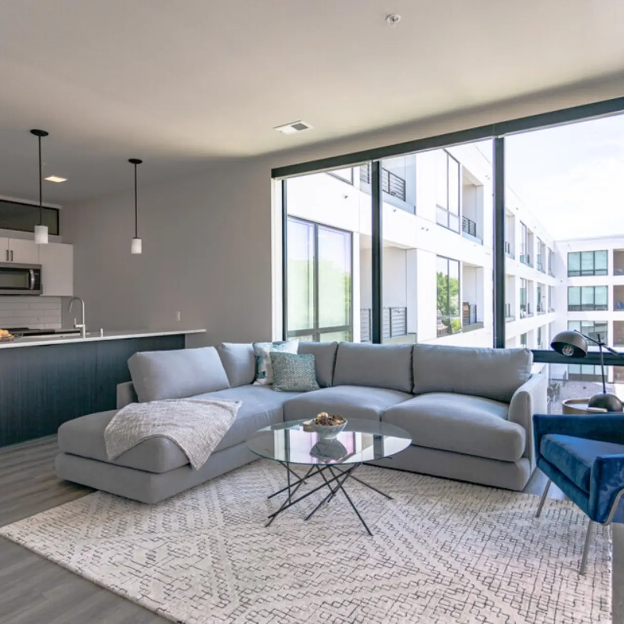 A modern living room featuring a large gray sectional sofa, a glass coffee table, and large windows allowing natural light. The room includes an adjacent kitchen with stainless steel appliances and a sleek design.