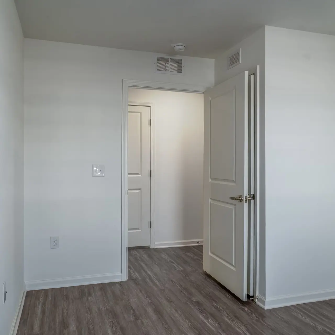 An empty room with light beige walls and laminate flooring. Two doors can be seen; one is in the foreground on the right, and another door is visible in the back to the left. Bright natural light filters in through an unseen window.