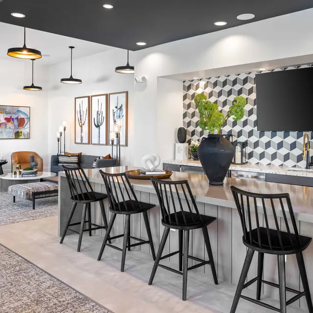 A modern lounge area featuring a bar with black stools, geometric wall decor, a television, and cozy seating arrangements with two chairs. The decor includes pendant lighting and various indoor plants.