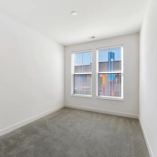 Empty Bedroom with Windows A vacant bedroom with gray carpet and light colored walls, featuring two windows with blinds.