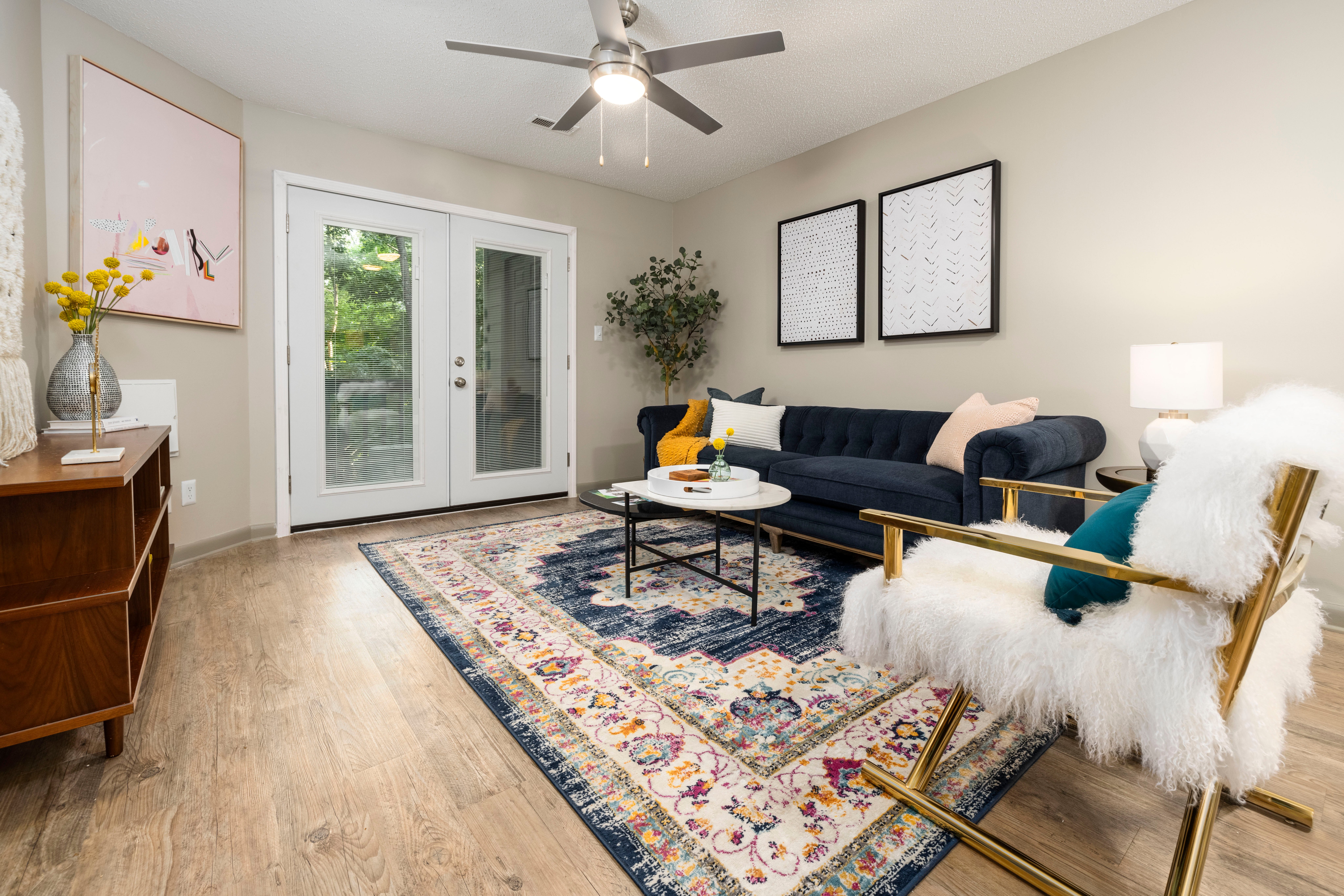 A modern living room featuring a navy blue couch, colorful decorative pillows, a round coffee table, and an elegant chair with a fluffy white seat. The room has wooden flooring, large glass doors leading outside, and small plants. Two framed artworks hang on the wall.