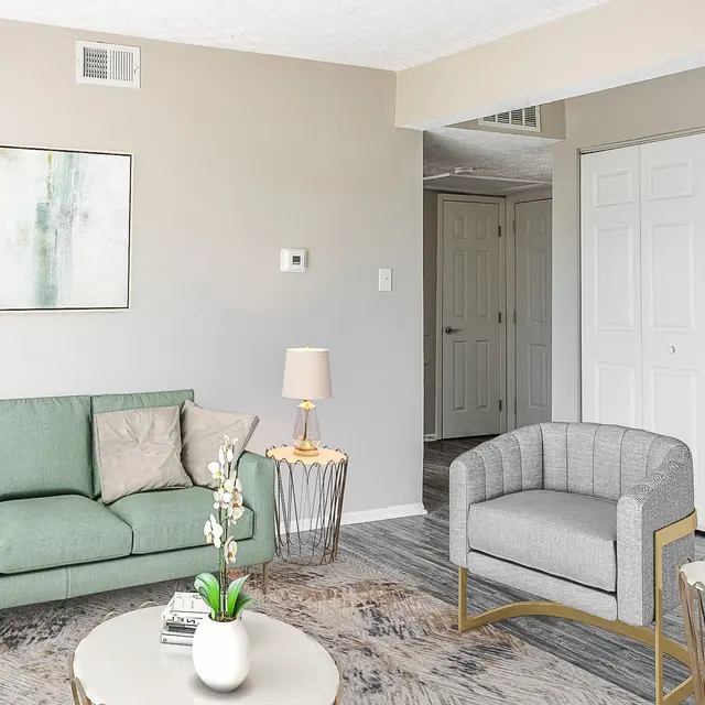 The Hideaway -  A modern living room with a light-colored sofa and gray armchair, featuring a round coffee table and a decorative plant. A wall art piece hangs above the sofa, and a lamp sits next to it, with a closet door visible in the background.