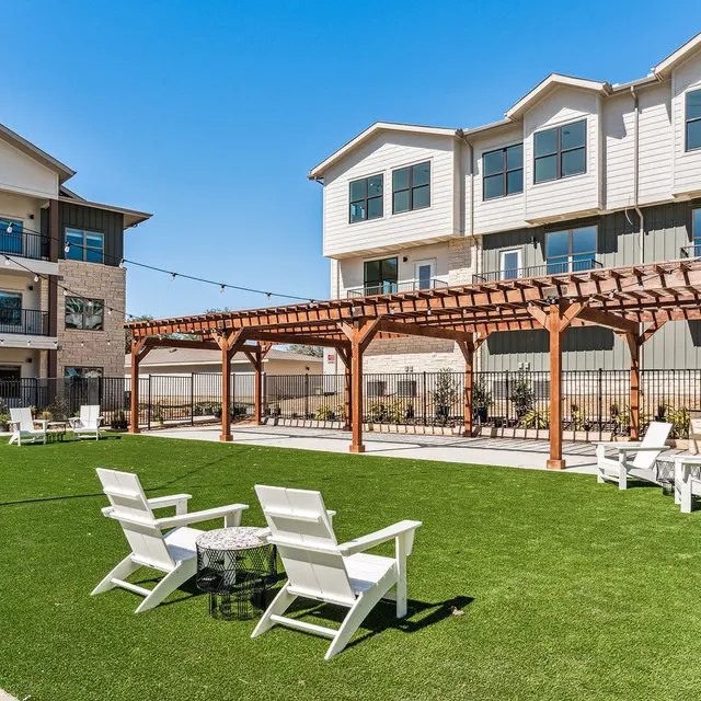 A landscaped outdoor area with green grass, white lounge chairs, a wooden pergola, and modern apartment buildings in the background under a clear blue sky.