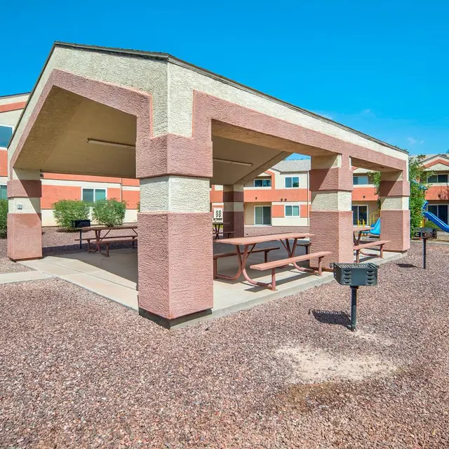 An outdoor pavilion area with several picnic tables surrounded by landscaping and residential buildings, under a clear blue sky.