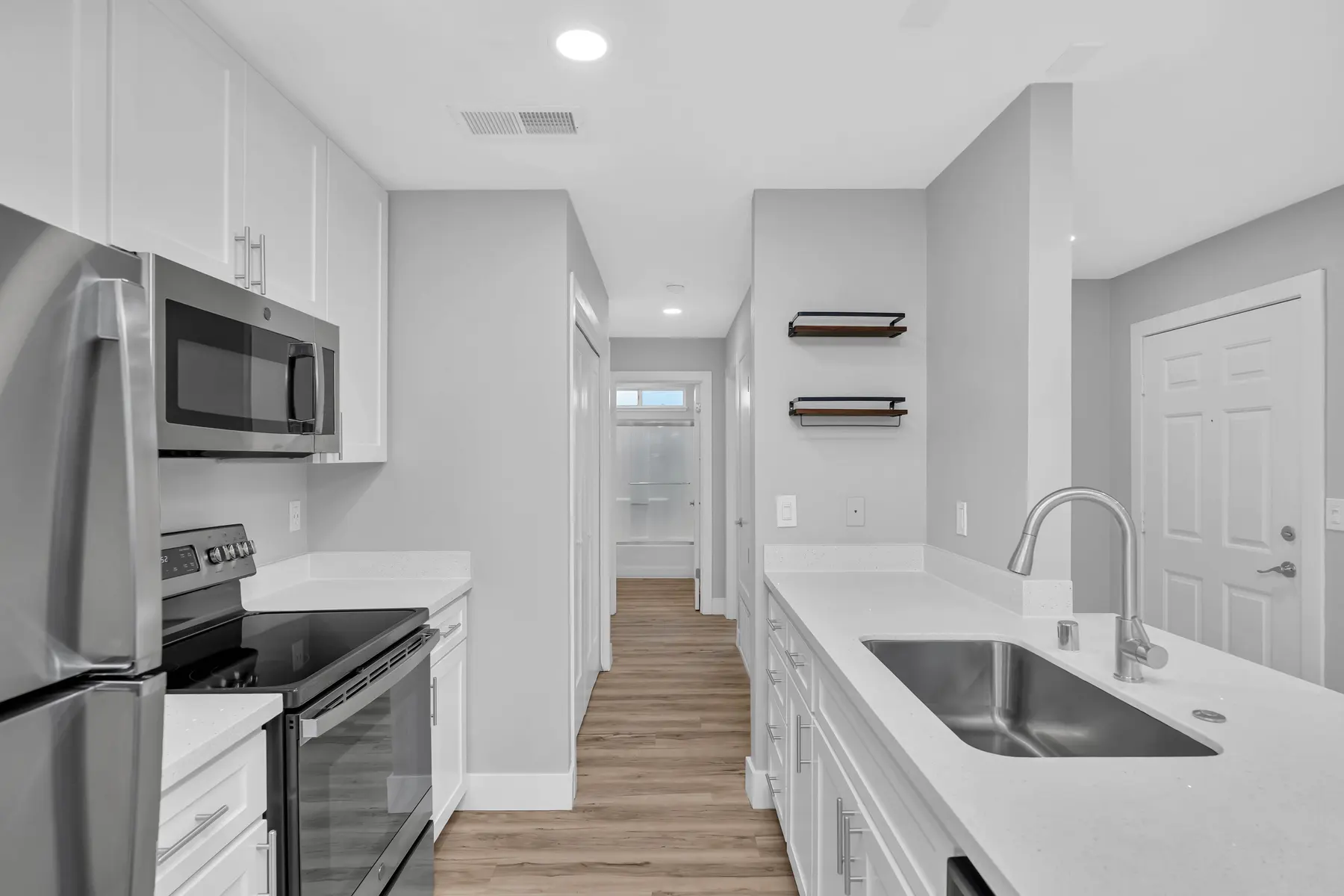 A modern kitchen featuring stainless steel appliances, white cabinetry, and a white countertop. The kitchen layout is open, showcasing a view of a hallway leading to a bright room. The floor is made of light wood and there are minimal decorative elements on the walls.