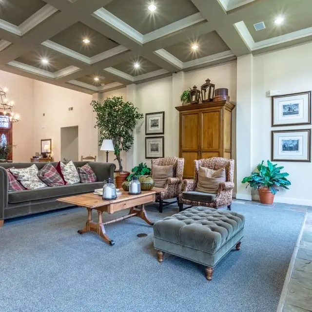 Cozy Living Room Interior A cozy living room with a plush sofa, armchairs, a coffee table, and decorative plants. The ceiling features recessed lighting, and the walls have framed pictures. There is a fireplace and a wooden cabinet in the background.