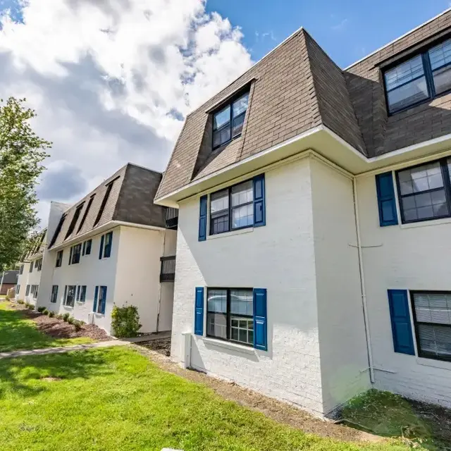 Exterior view of a multi-unit apartment building with white walls and blue shutters, set against a partly cloudy sky.