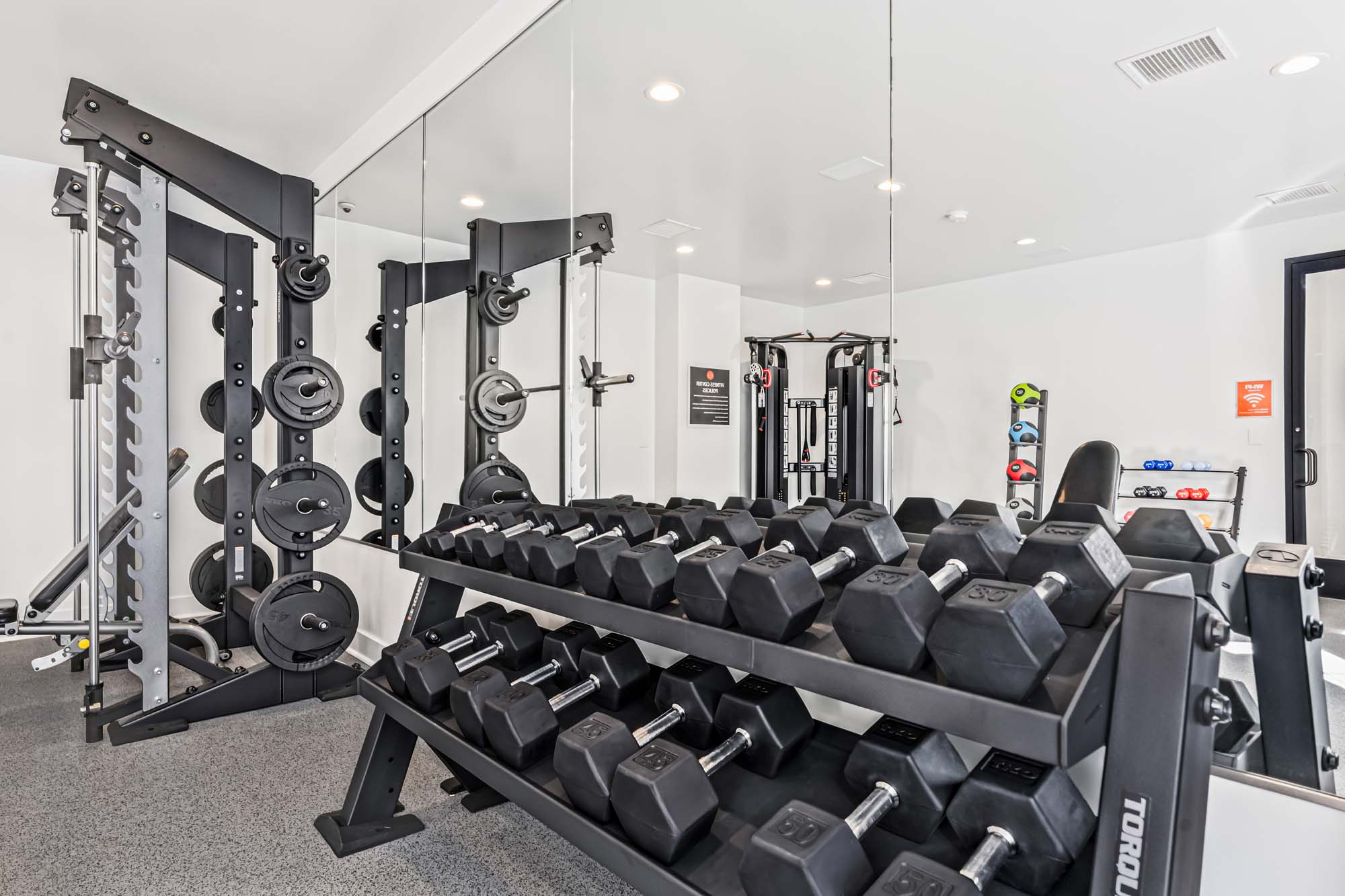 A modern gym area featuring a rack of dumbbells, adjustable weight equipment, and mirrors on the wall.
