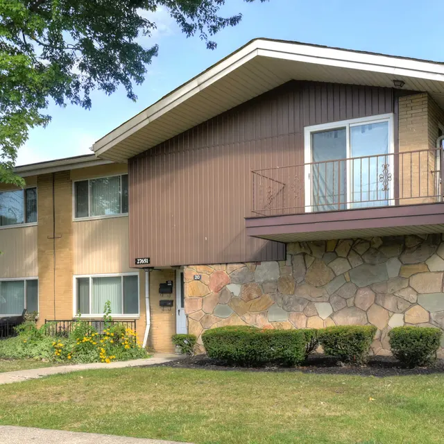 Exterior view of a two-story apartment building with a mix of brick and stone facade, surrounded by greenery and a walking path.