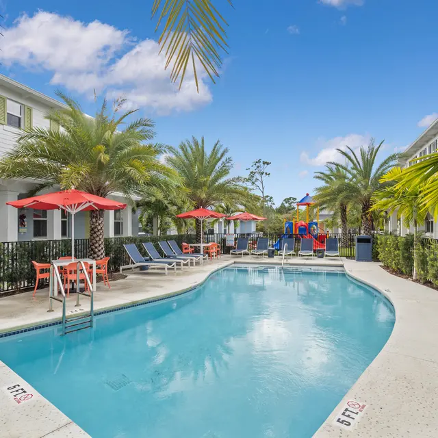 A swimming pool surrounded by palm trees and lounge chairs, with colorful umbrellas and a small playground in the background.