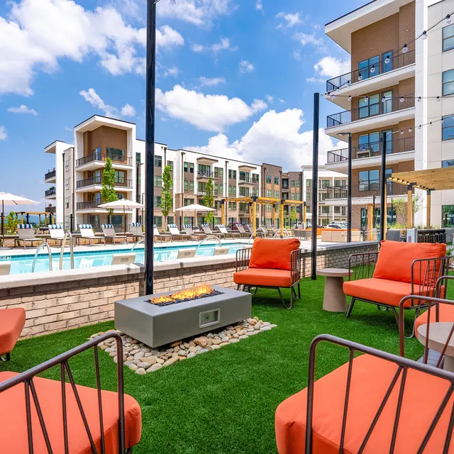 A modern apartment complex featuring a luxurious outdoor pool area, surrounded by lounge chairs and a vibrant green lawn. The buildings are contemporary with multiple balconies and stylish architecture under a clear blue sky.