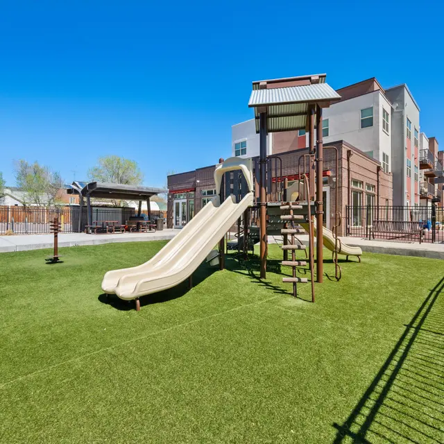 A playground area featuring a slide, climbing structure, and green synthetic turf at Baker School Apartments in Berkley, Colorado.