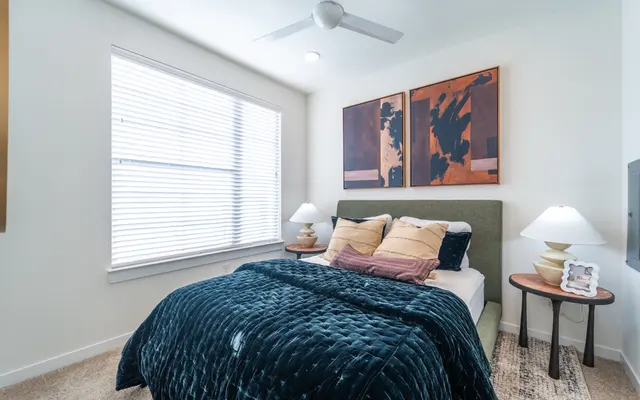 Modern Cozy Bedroom A cozy bedroom with a green upholstered bed, adorned with a plush navy blue blanket and decorative pillows. There are two bedside lamps and artwork on the wall above the bed. Natural light streams in through a window with white blinds.