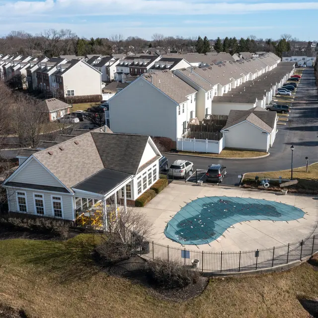 Aerial view of a residential community featuring a swimming pool, surrounded by houses and parked cars, with clear blue skies overhead.