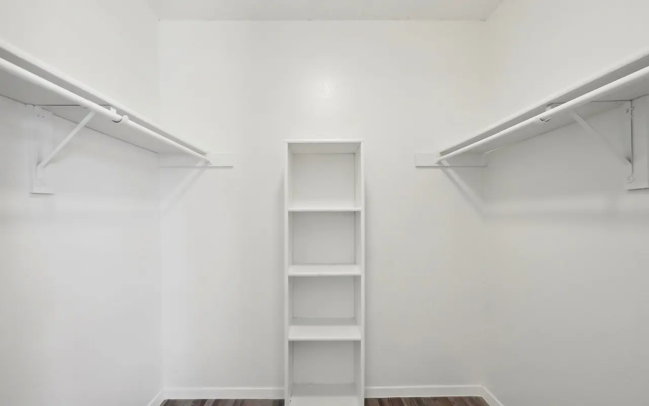 An empty closet with two hanging racks on either side and a white shelving unit in the center, featuring light-colored wooden flooring.