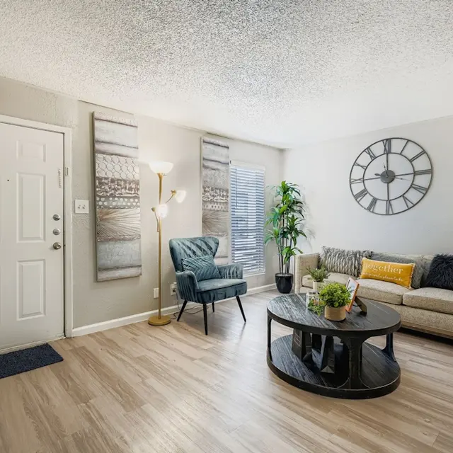 Modern Living Room Interior A modern living room featuring a beige couch with colorful cushions, a dark circular coffee table, green potted plants, and decorative wall art. A round wall clock is mounted above the couch, and the entrance shows a light-colored door.