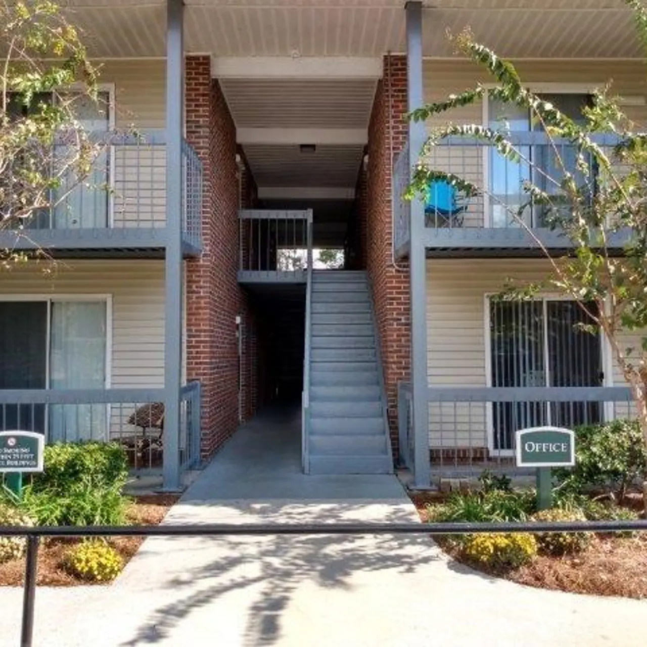 View of an apartment complex showing two floors with balconies, a central staircase, and greenery in front.