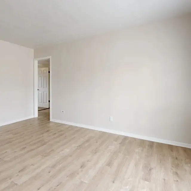 Spacious empty room with light wooden flooring and neutral-colored walls, with a window providing natural light.
