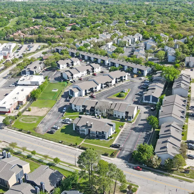 Aerial view of a residential area featuring multiple apartment complexes, parking spaces, and green spaces surrounded by trees.