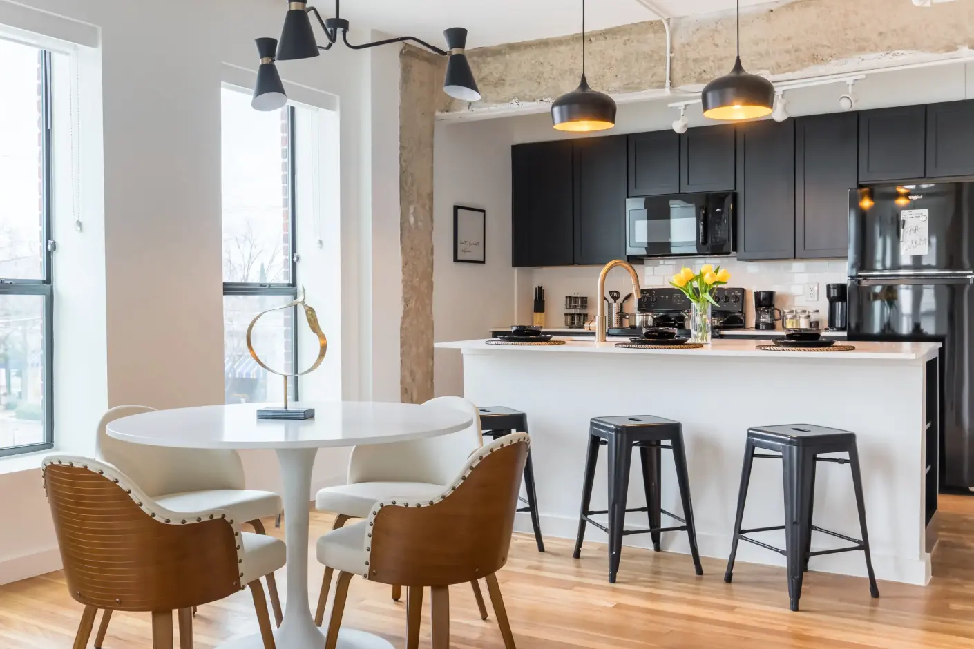 A modern kitchen and dining area featuring a round white table with four chairs, a black kitchen with stainless steel appliances, and wooden flooring.