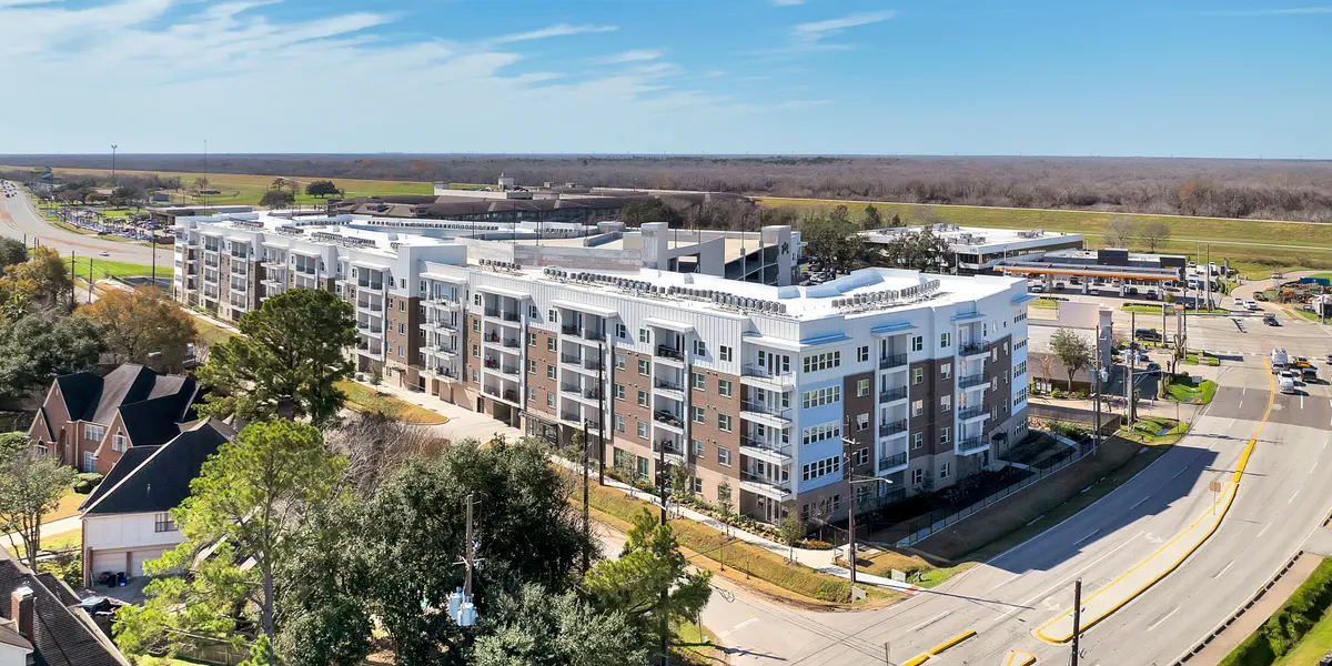Aerial view of a modern apartment building alongside a road, surrounded by trees and open fields in the background under a clear blue sky.