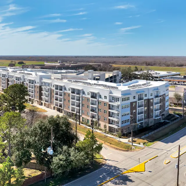 Aerial view of a modern apartment building alongside a road, surrounded by trees and open fields in the background under a clear blue sky.