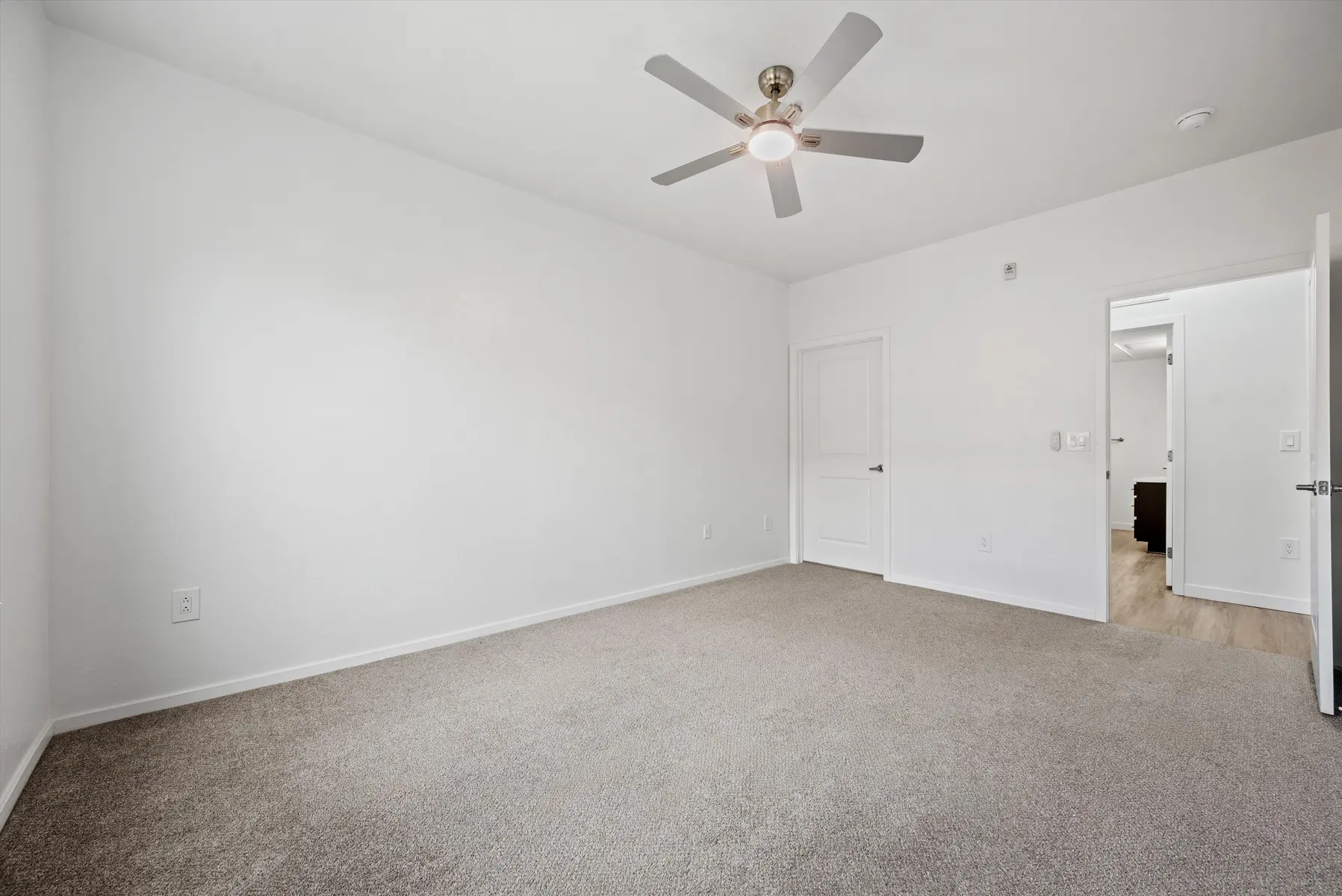 An empty bedroom featuring beige carpet, a ceiling fan, and white walls with a door leading to another room.