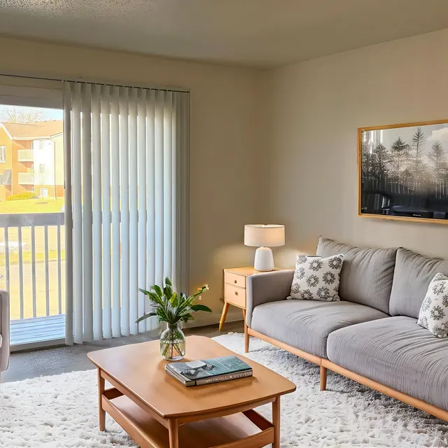 A cozy living room featuring a gray sofa with decorative pillows, a wooden coffee table, and a comfortable armchair. Natural light fills the space through sliding glass doors, revealing a view of the outdoors. A framed landscape photograph is mounted on the wall, and a small vase with flowers sits on the coffee table.