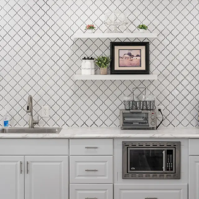 A modern kitchen with white cabinetry and a patterned backsplash. There are open shelves with decorative items, including a framed photograph and potted plants. The countertop is light-colored and a microwave is built into the cabinetry.